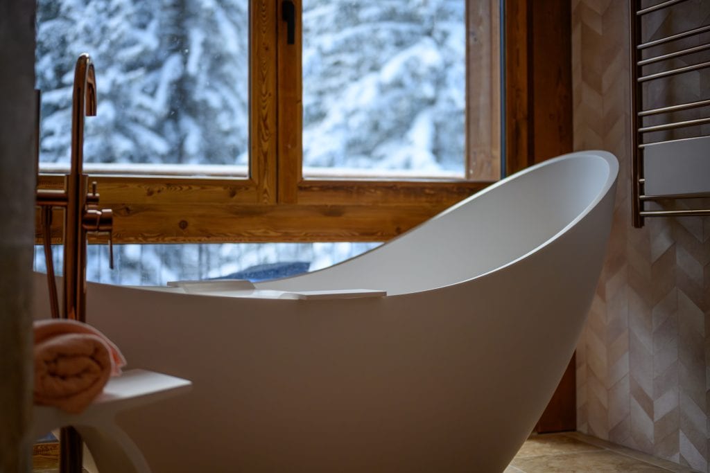Close-up of a freestanding bathtub with snowy forest view through large windows.
