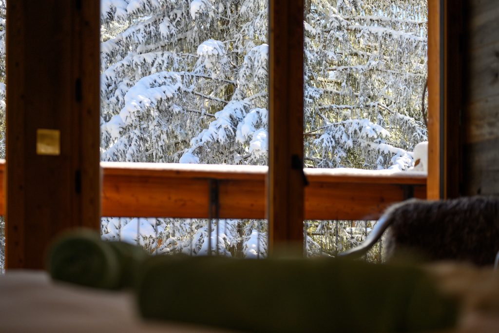 Snowy pine trees seen through a bedroom window and balcony rail.