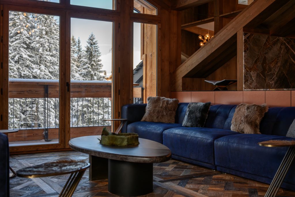Close-up of blue velvet sofa with fur cushions beside large windows and forest views.