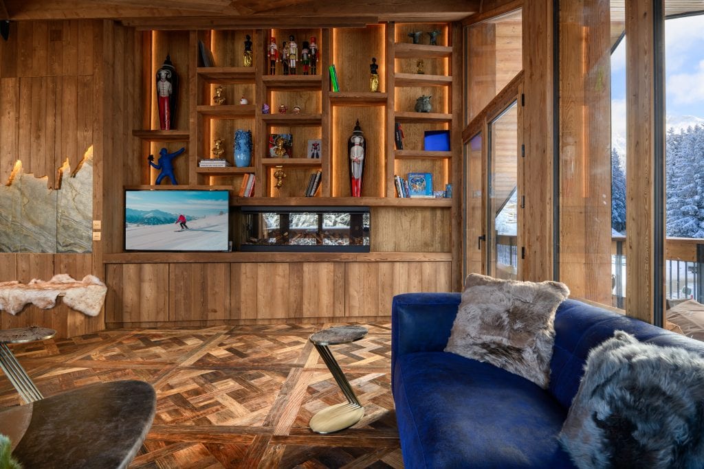 Living room with custom wooden bookshelf, blue velvet sofa, and large windows overlooking snowy forest.